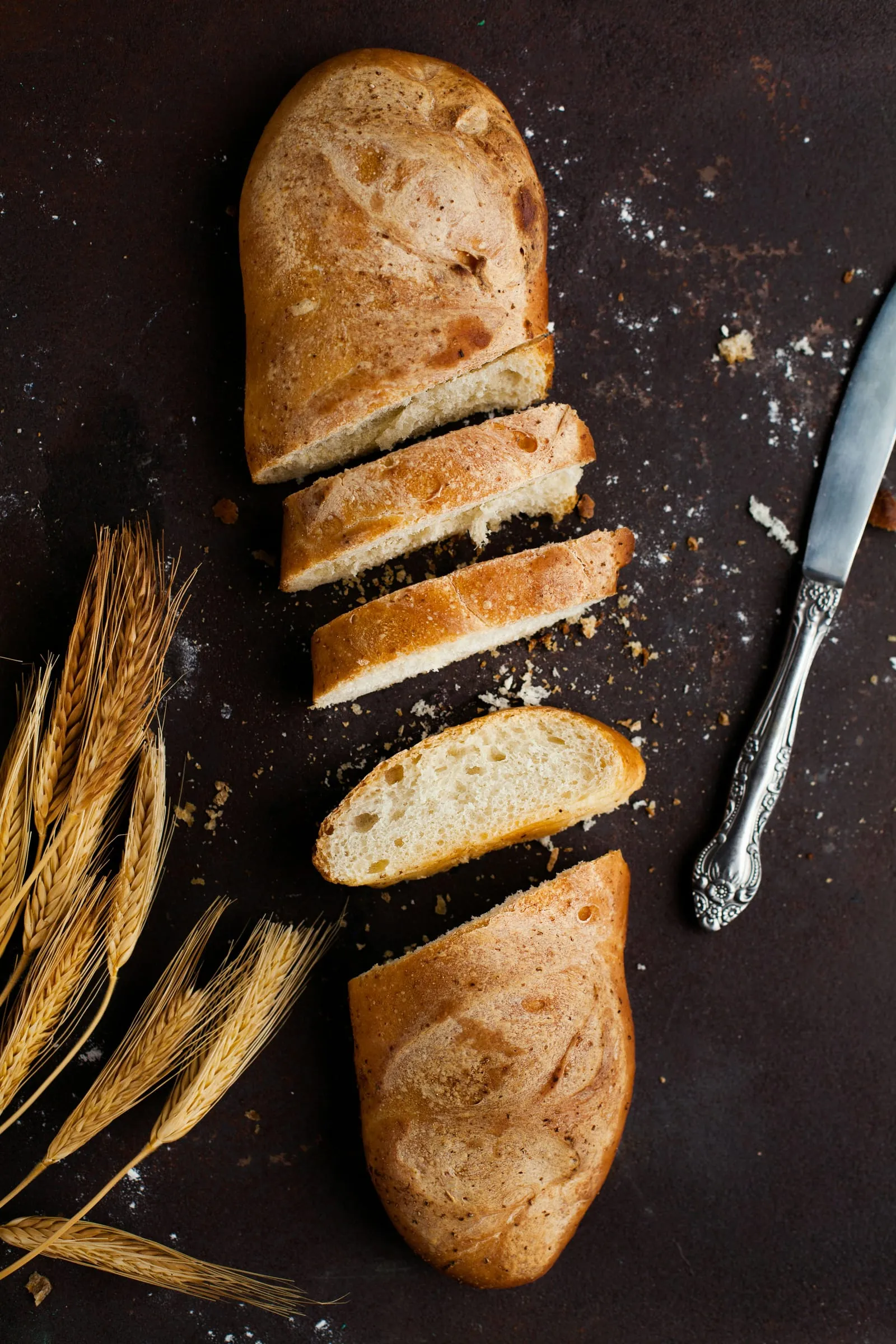 Gesneden wit brood met kruimels, tarwearen en een tafelmes op een donkere ondergrond
