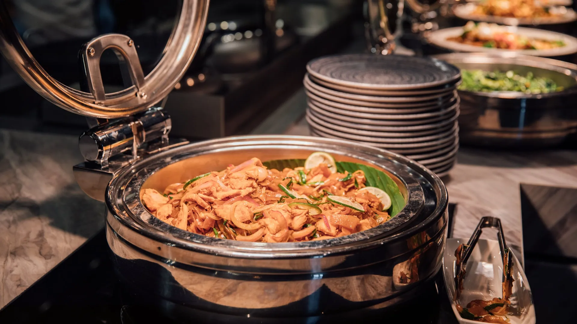 Open warmhoudschaal met kip, ui en limoen op een warm buffet, borden erachter