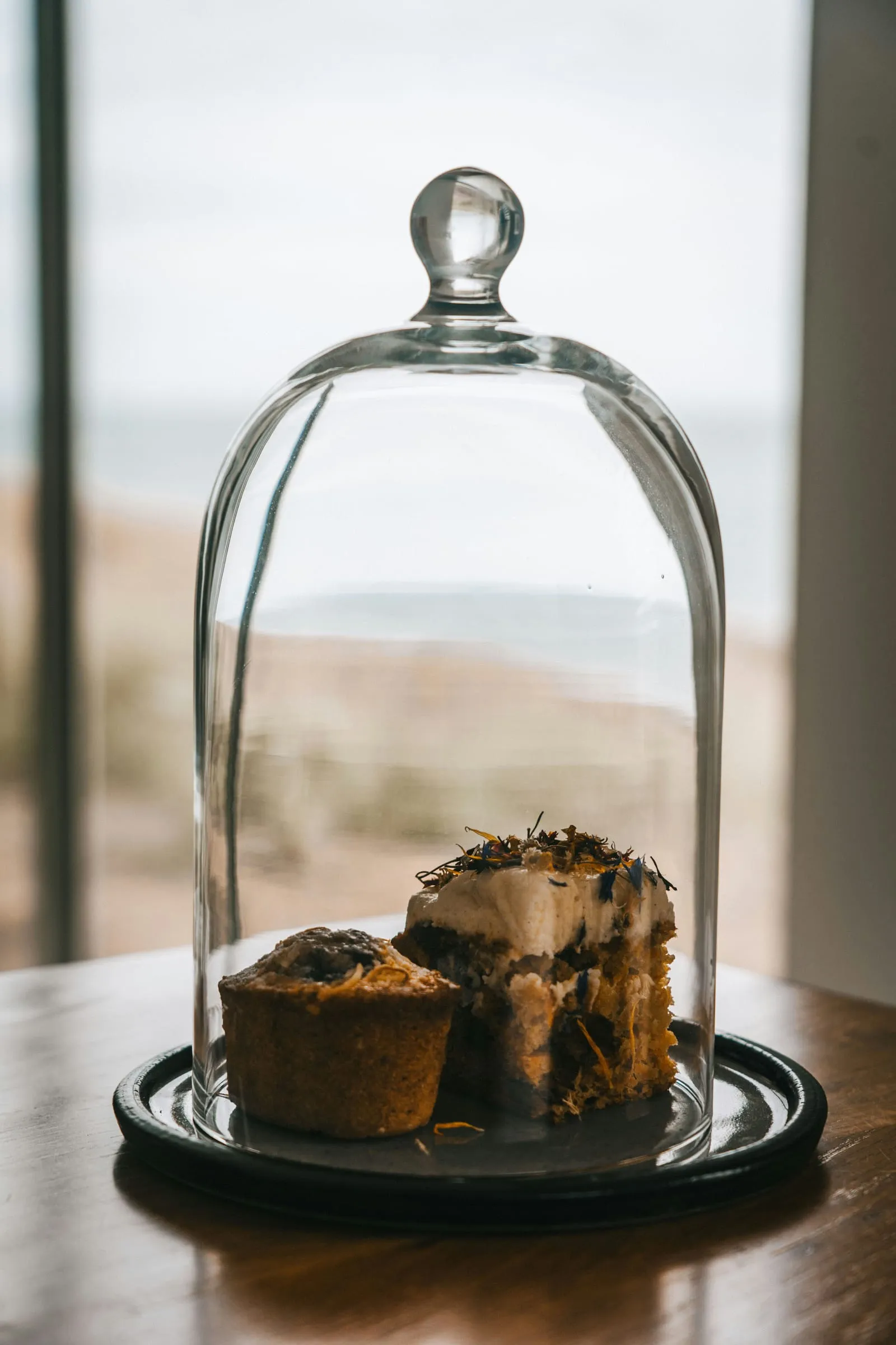 Een stuk taart en een muffin onder een glazen stolp bij het raam