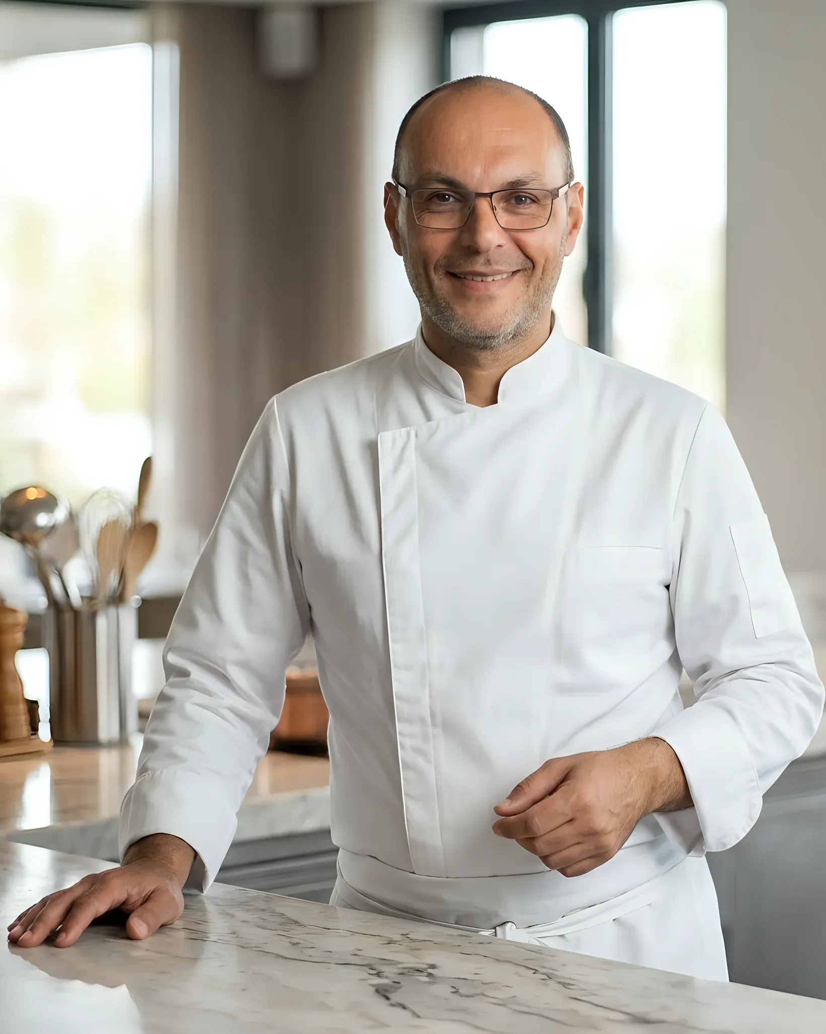 David Nagib in witte koksbuis, lachend aan een marmeren aanrecht in een lichte keuken