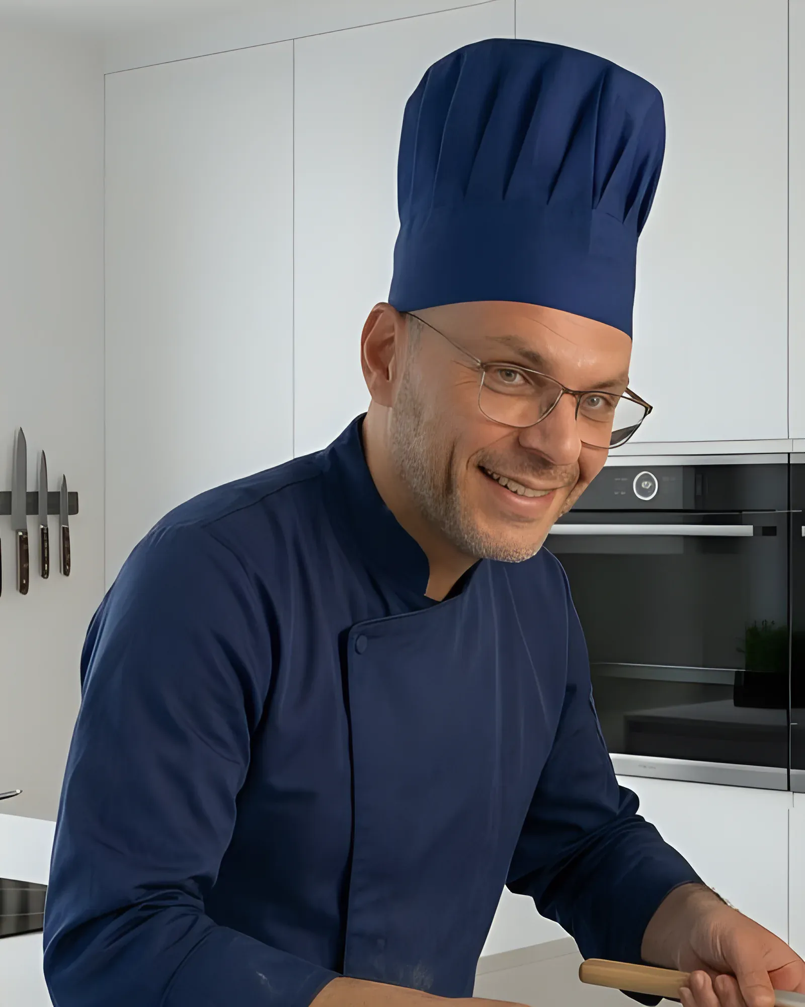 David Nagib in donkerblauwe koksbuis en koksmuts, lachend in de keuken