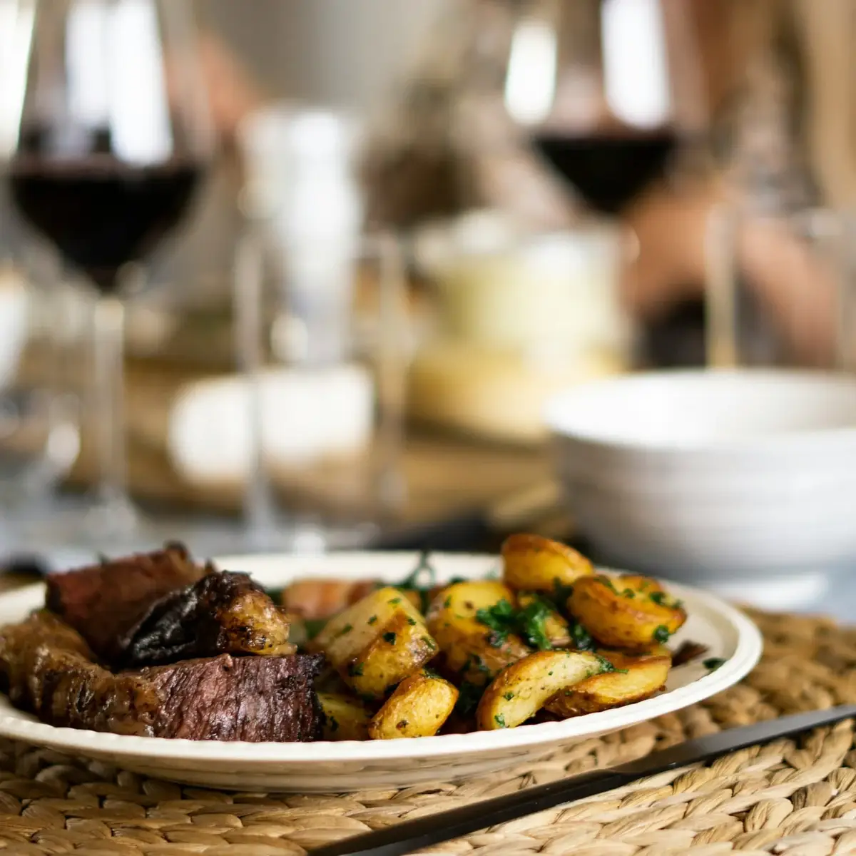 Diner aan tafel: vlees met gebakken aardappeltjes, met wijnglazen op de achtergrond