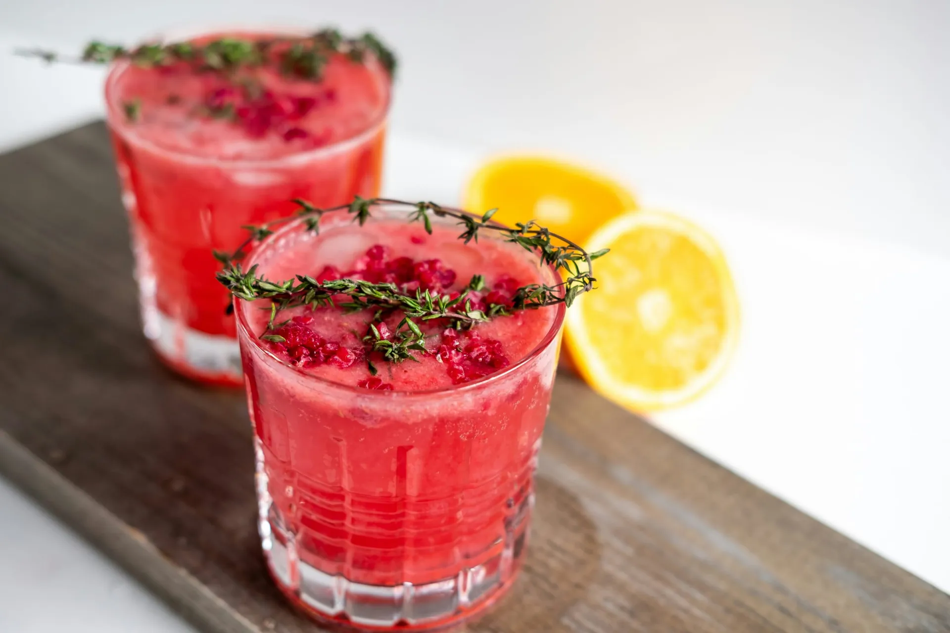 Twee glazen rood fruitdrankje met frambozen en tijm, naast een gehalveerde sinaasappel