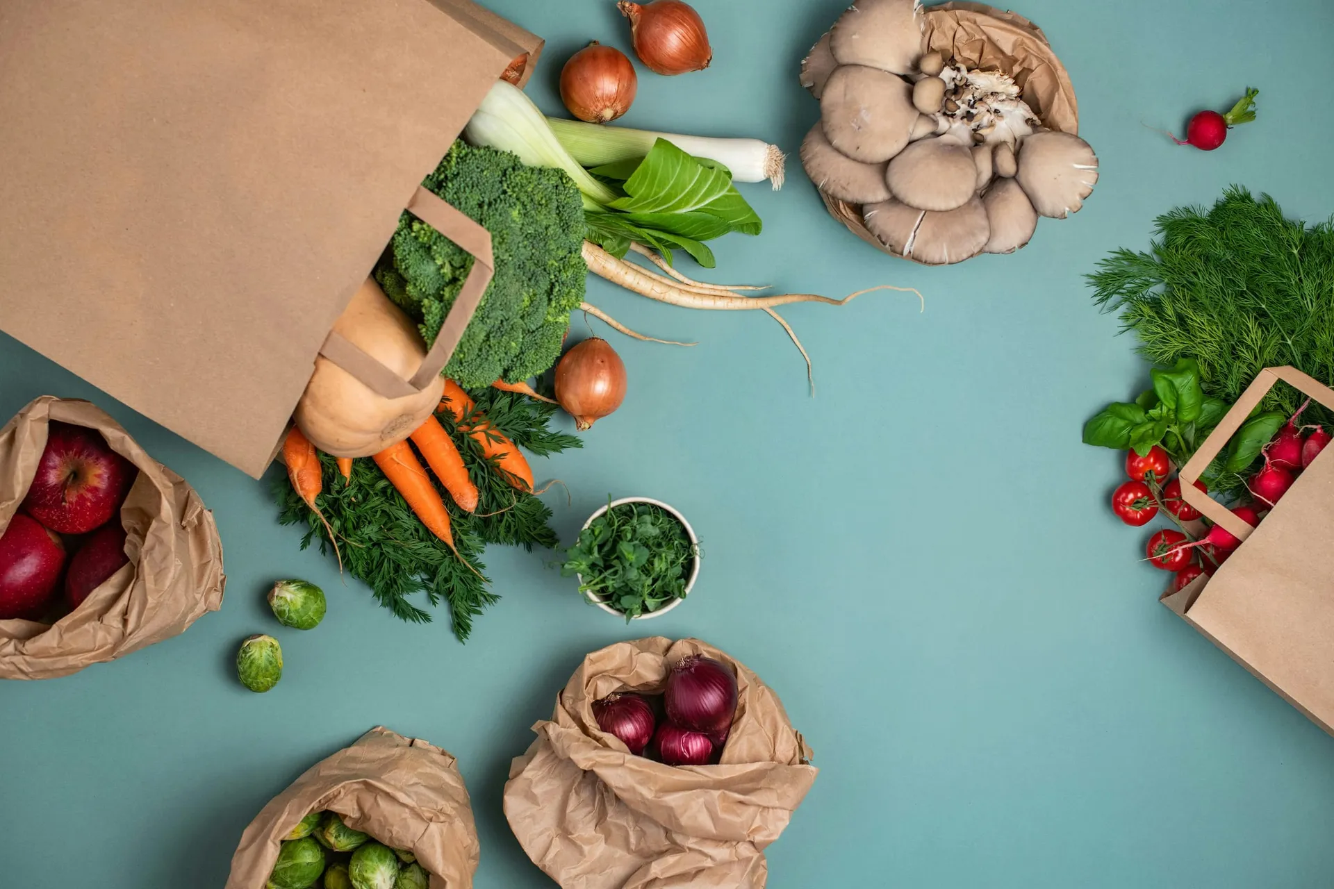 Papieren zakken met wortels, broccoli, uien, oesterzwammen, radijs, spruitjes en appels op een blauwgroene ondergrond
