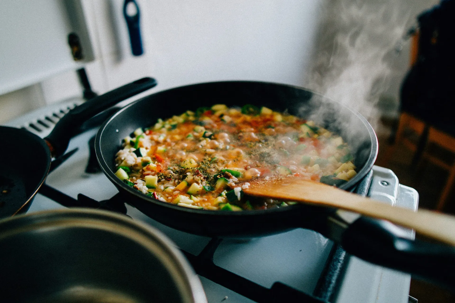 Een koekenpan met rijst, courgette en paprika die stoomt op het gasfornuis, met houten spatel