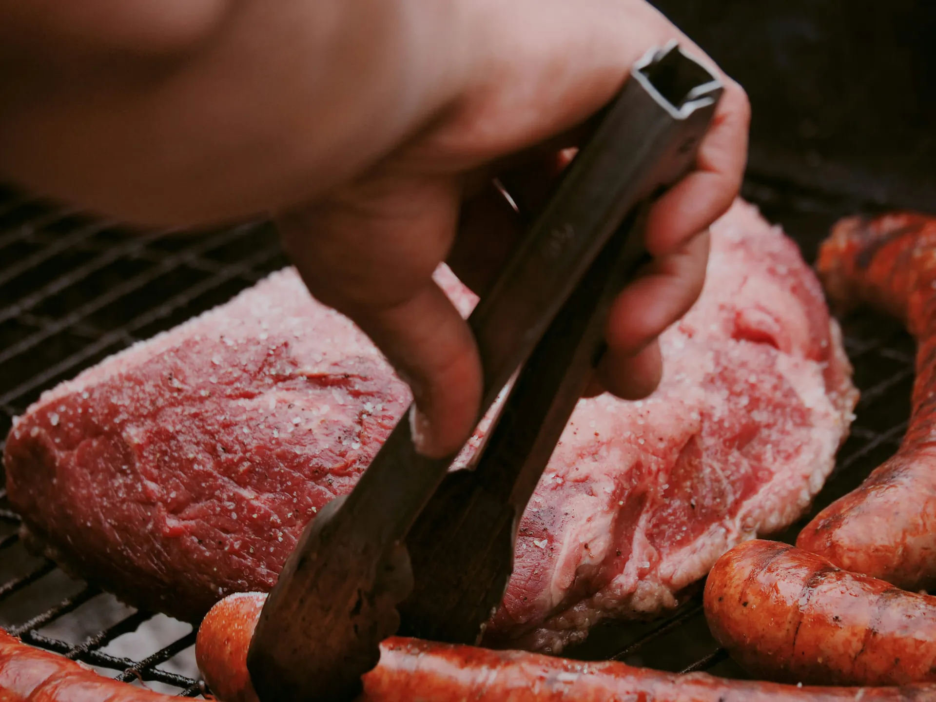 Een hand met een barbecuetang boven een rauwe gezouten steak en gegrilde worsten